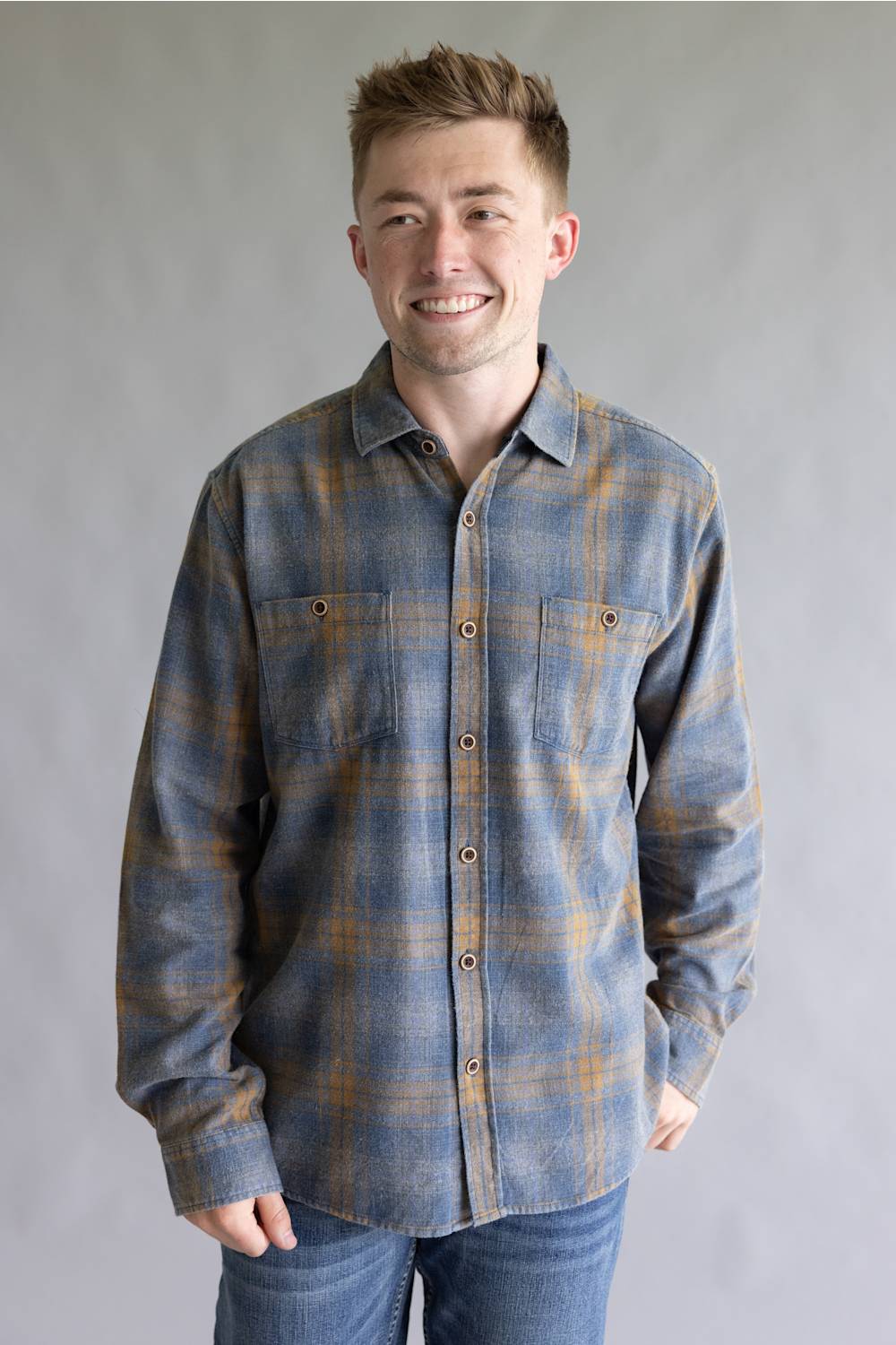A smiling man wearing a long-sleeve plaid button-up shirt in blue and brown tones, paired with blue jeans, standing against a neutral gray studio background.