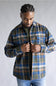A man wearing a blue, green, and tan plaid button-up flannel shirt over a black t-shirt and blue jeans, standing against a neutral gray studio background.