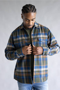 A man wearing a blue, green, and tan plaid button-up flannel shirt over a black t-shirt and blue jeans, standing against a neutral gray studio background.