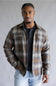 A man wearing a brown and beige plaid button-up shirt over a black henley t-shirt and light wash jeans, standing against a neutral gray studio background.