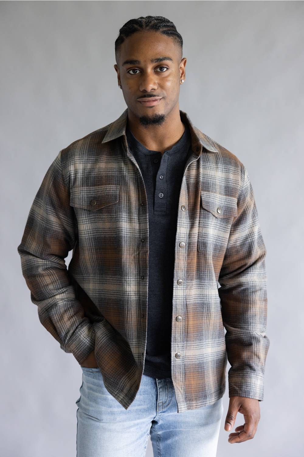 A man wearing a brown and beige plaid button-up shirt over a black henley t-shirt and light wash jeans, standing against a neutral gray studio background.