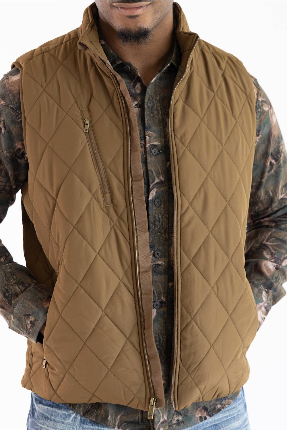 A man wearing a tan diamond-quilted zip-front vest over a camouflage long-sleeve shirt and blue jeans, shown from the lower face to the upper thighs on a white background.