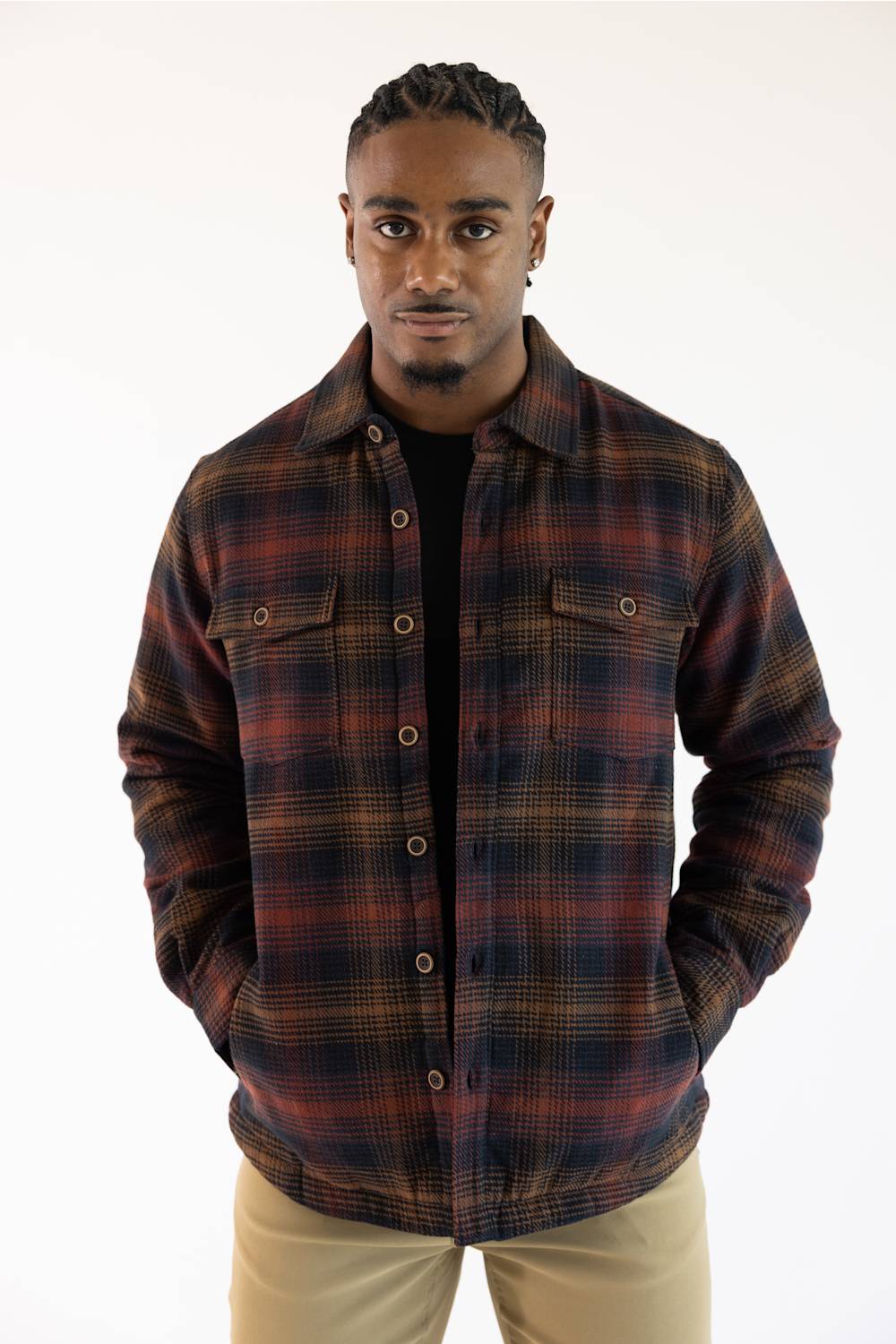 A man wearing a dark brown and red plaid button-up flannel shirt over a black t-shirt and khaki pants, standing against a plain white background.