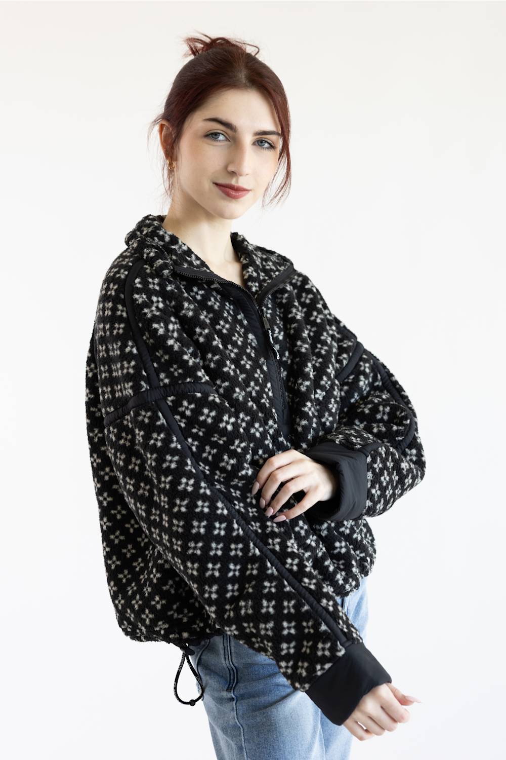 A woman wearing a black and white patterned zip-up hoodie jacket with black ribbed cuffs and hem, paired with light blue jeans, photographed against a plain white studio background.
