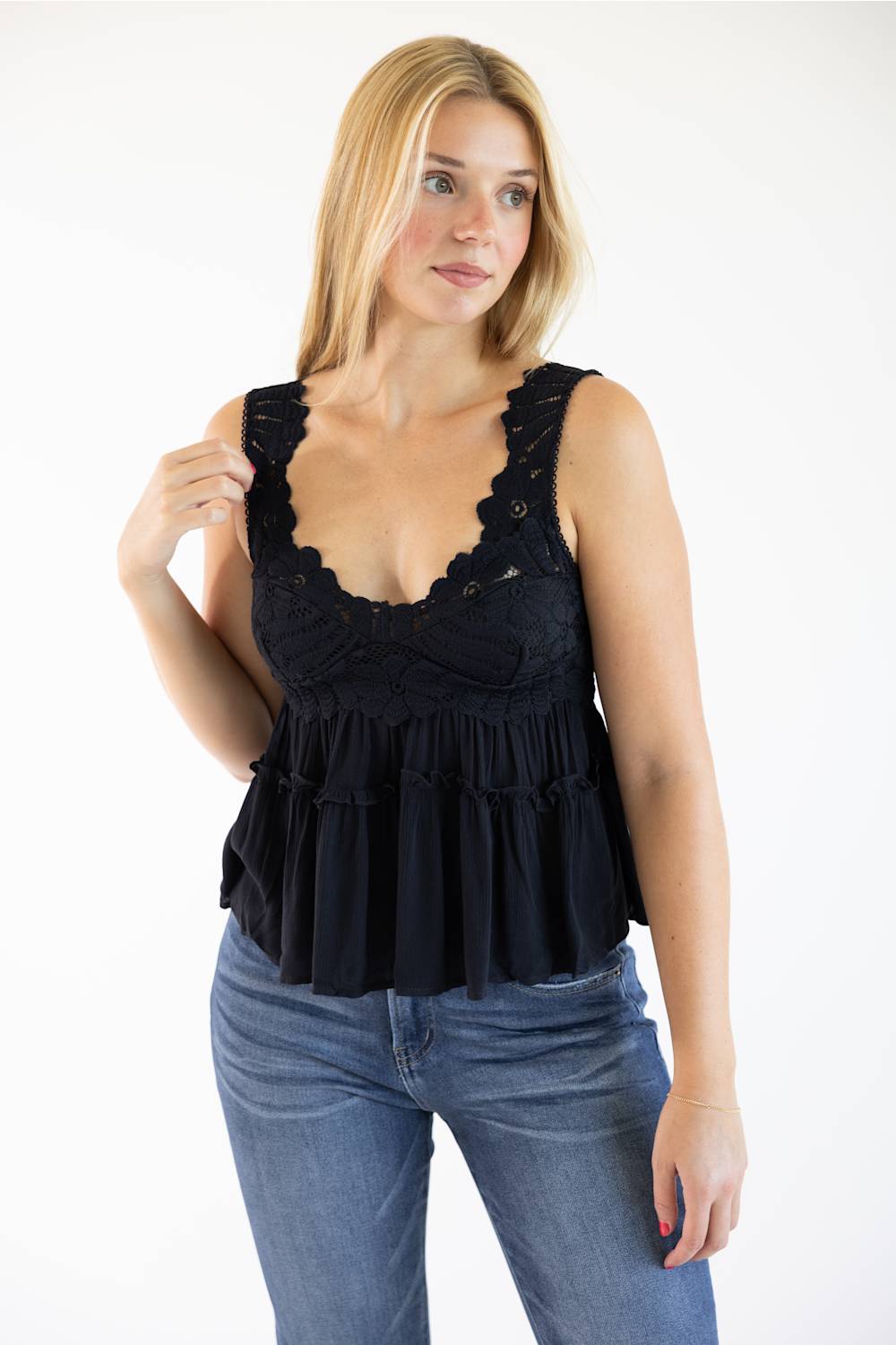 A woman modeling a black sleeveless ruffled top with scalloped lace trim and a tiered peplum, paired with blue jeans, against a plain white background.