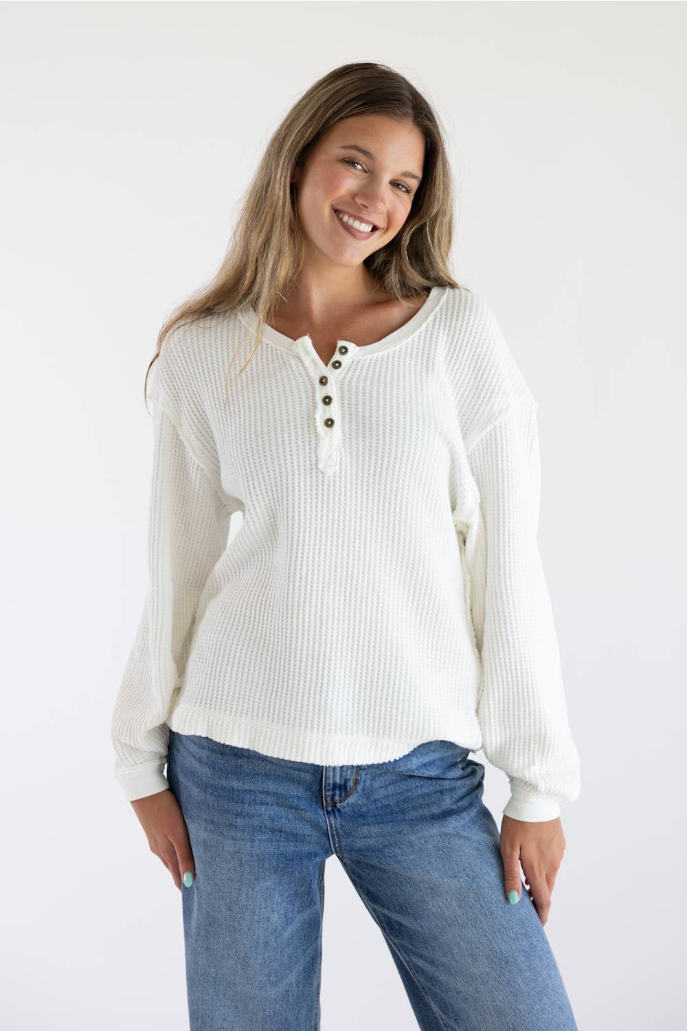 A woman wearing a white ribbed long-sleeve henley sweater with brown buttons, paired with blue jeans, against a plain white studio background.