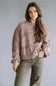 A woman modeling a dusty rose open-knit sweater with balloon sleeves and olive green pants, standing against a solid gray studio background.
