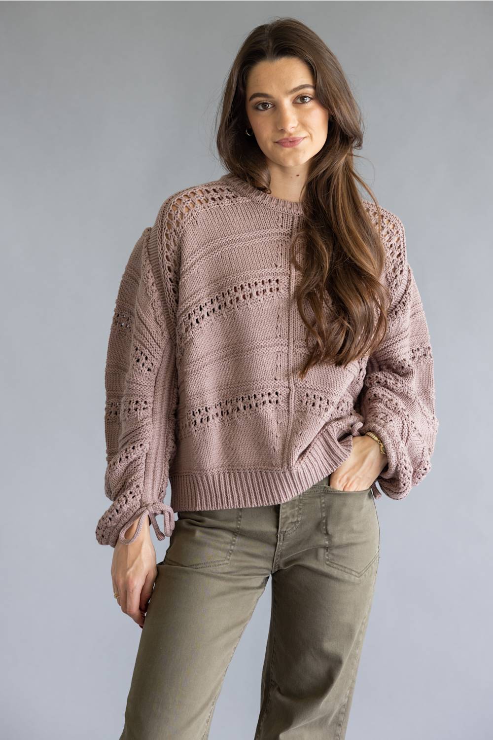 A woman modeling a dusty rose open-knit sweater with balloon sleeves and olive green pants, standing against a solid gray studio background.