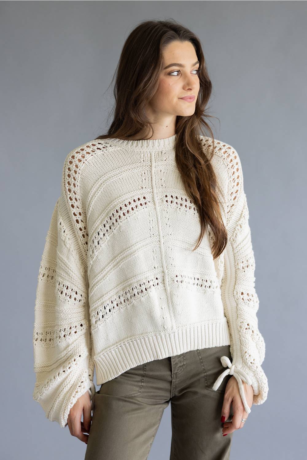 A woman wearing a cream-colored, open-knit crewneck sweater with balloon sleeves and olive green pants, standing against a solid gray studio background.