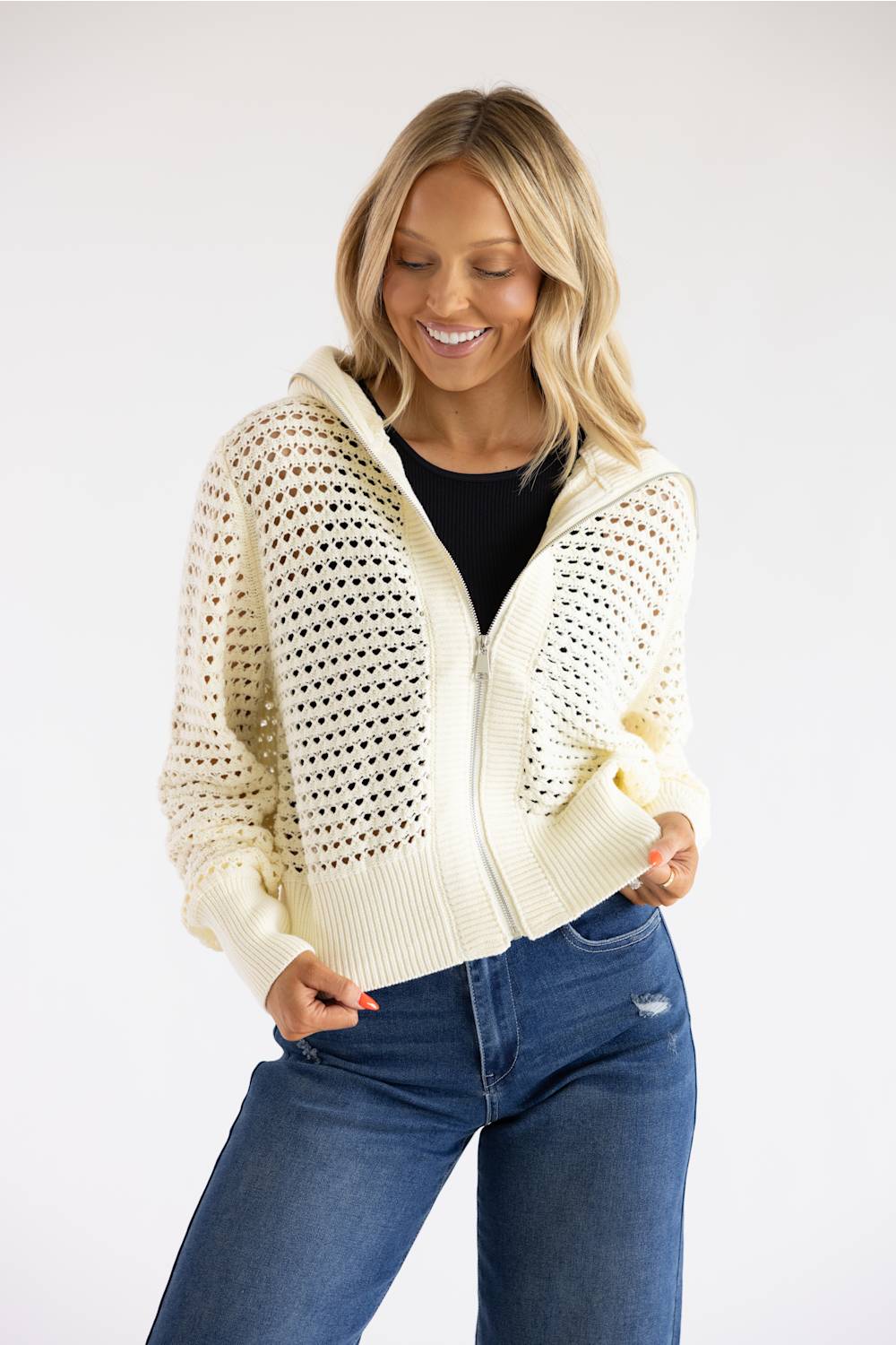A woman wearing a cream-colored open-knit zip-up hoodie with a hood, styled over a black top and blue jeans, posing against a plain white studio background.