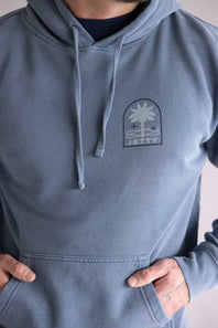 Close up of a blue hoodie with a palm tree logo and 'Jetty' text on a neutral background. The hoodie is the Jetty Orbit Grove Hoodie for Men in Pigment Slate