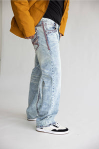 A person wearing light-wash blue denim jeans with decorative red and black contrast stitching, styled with a black top, a mustard yellow jacket, and black-and-white sneakers against a plain light gray studio background.