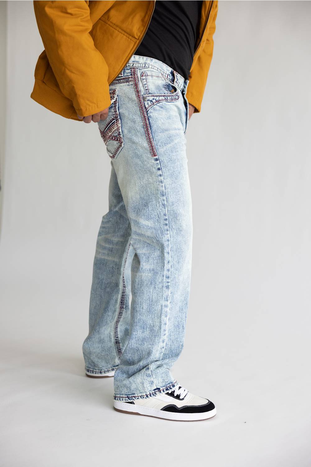 A person wearing light-wash blue denim jeans with decorative red and black contrast stitching, styled with a black top, a mustard yellow jacket, and black-and-white sneakers against a plain light gray studio background.