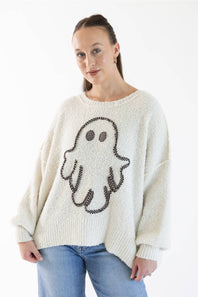 A woman wearing a cream-colored knit sweater with a large black and light orange gingham ghost design on the front, paired with light blue jeans, against a plain white background.