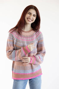 A smiling woman with long reddish-brown hair wearing a multicolor striped knit sweater with pink, peach, and light blue stripes, paired with blue jeans, against a plain white background.