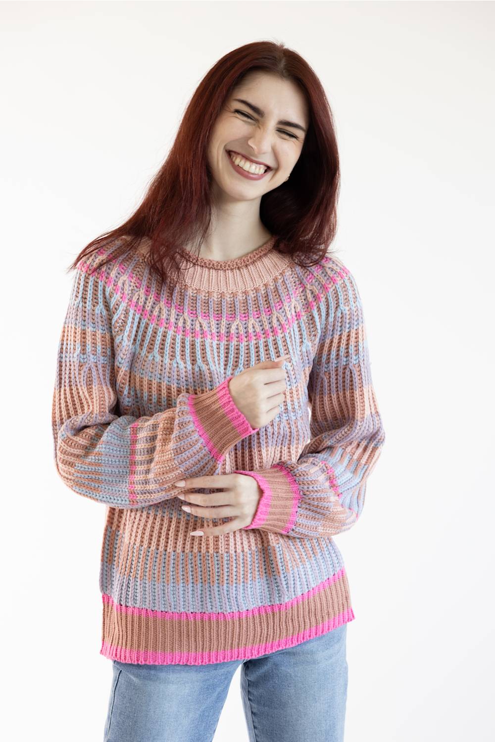 A smiling woman with long reddish-brown hair wearing a multicolor striped knit sweater with pink, peach, and light blue stripes, paired with blue jeans, against a plain white background.