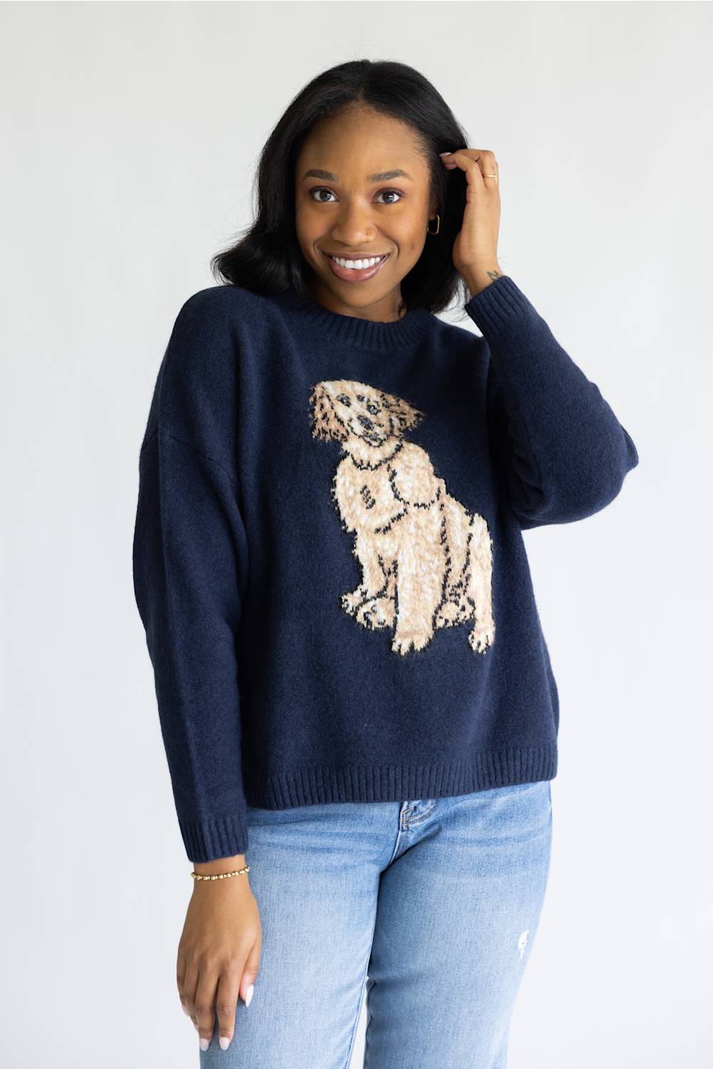 A woman wearing a navy blue knit sweater with a large beige and cream dog graphic on the front, paired with light blue jeans, against a plain white background.