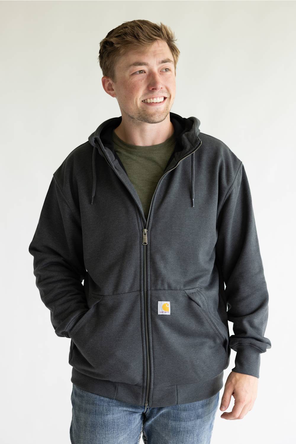 A man wearing a dark charcoal gray Carhartt zip-up hoodie over an olive green t-shirt and blue jeans, standing against a plain white background.
