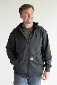 A man wearing a dark charcoal gray Carhartt zip-up hoodie over an olive green t-shirt and blue jeans, standing against a plain white background.