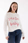 A woman wearing a white ribbed knit sweater with 'santa baby' written across the chest in red and white candy cane patterns, paired with dark blue jeans, against a plain white background.