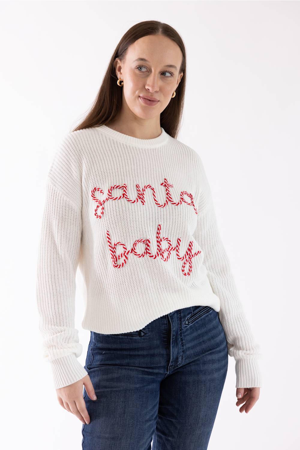 A woman wearing a white ribbed knit sweater with 'santa baby' written across the chest in red and white candy cane patterns, paired with dark blue jeans, against a plain white background.