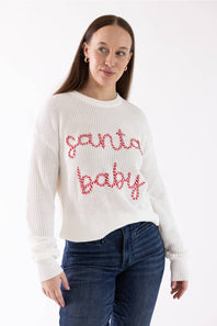 A woman wearing a white ribbed knit sweater with 'santa baby' written across the chest in red and white candy cane patterns, paired with dark blue jeans, against a plain white background.