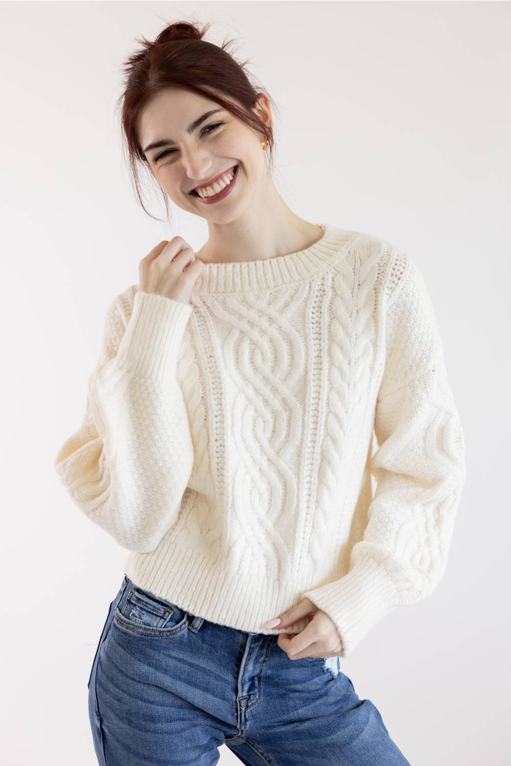A smiling woman wearing a cream cable-knit sweater and blue jeans, posing against a plain light background.