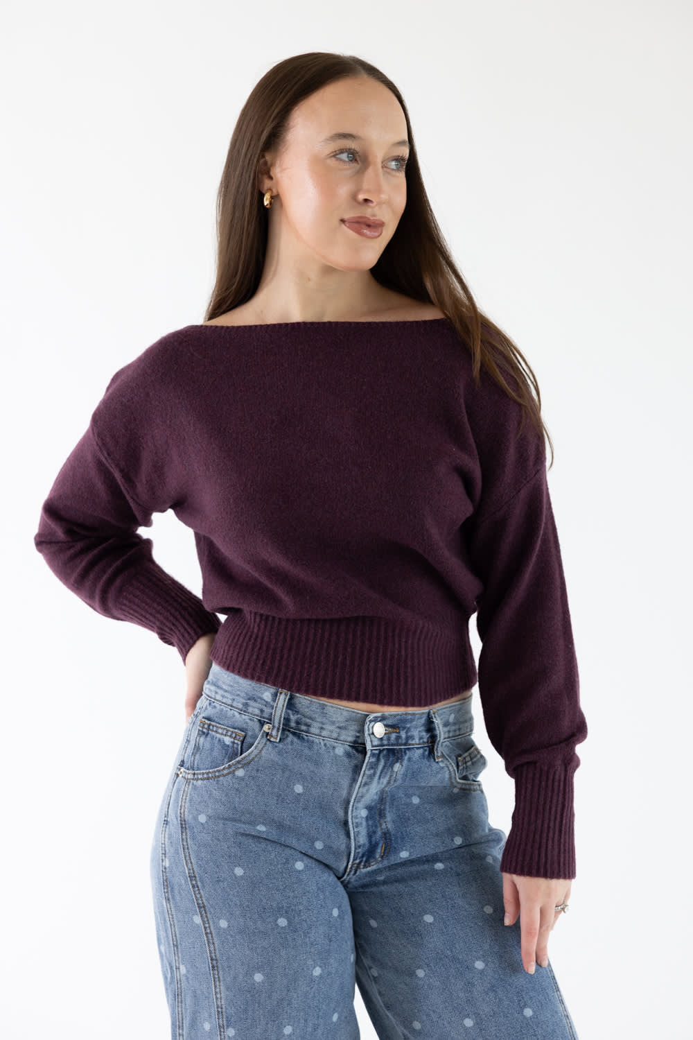 A woman modeling a dark plum purple off-the-shoulder knit sweater paired with blue jeans against a plain white studio background.