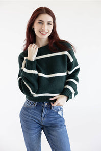 A woman with red hair wearing a dark green and white striped knit sweater and blue jeans, posing against a plain white studio background.