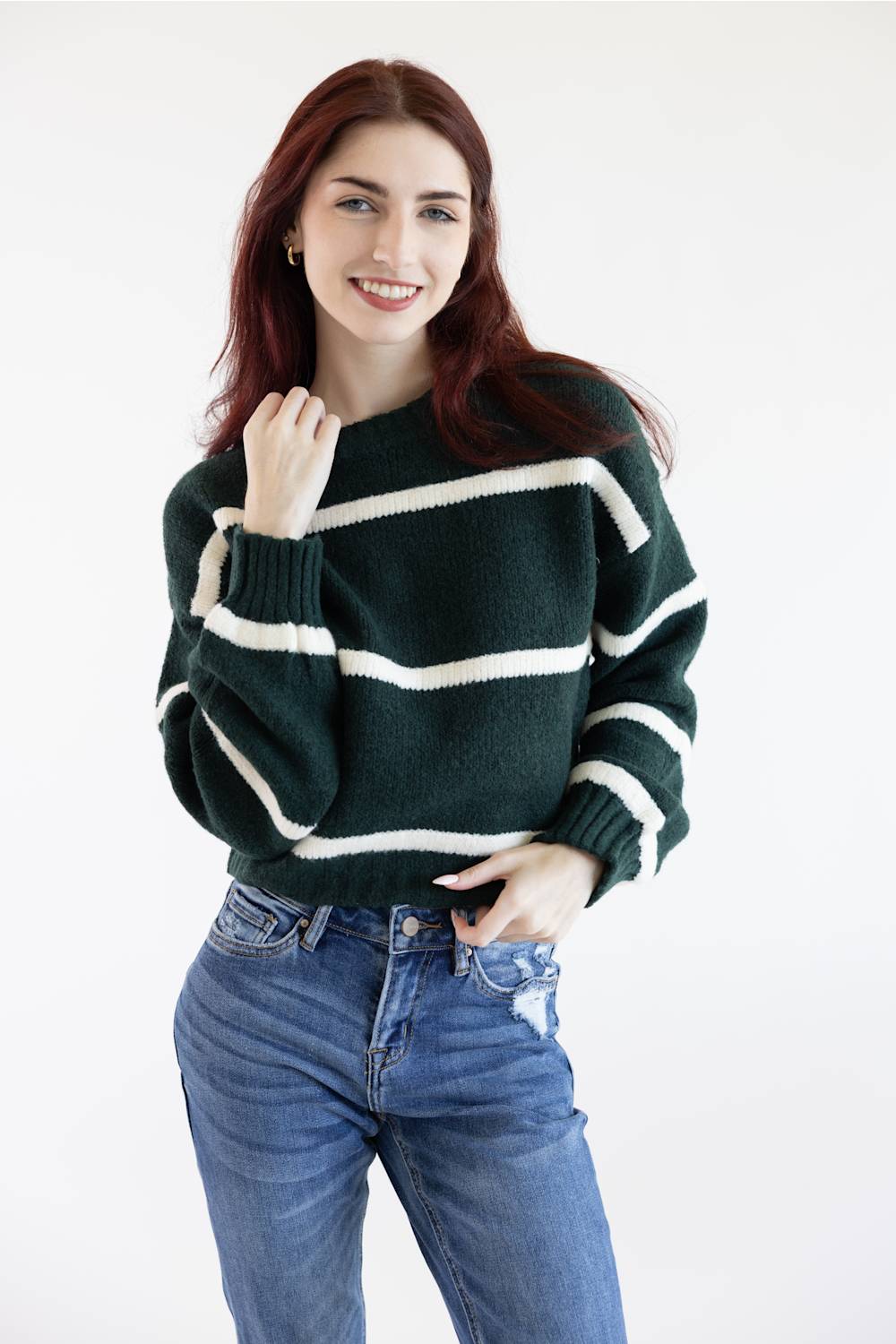 A woman with red hair wearing a dark green and white striped knit sweater and blue jeans, posing against a plain white studio background.