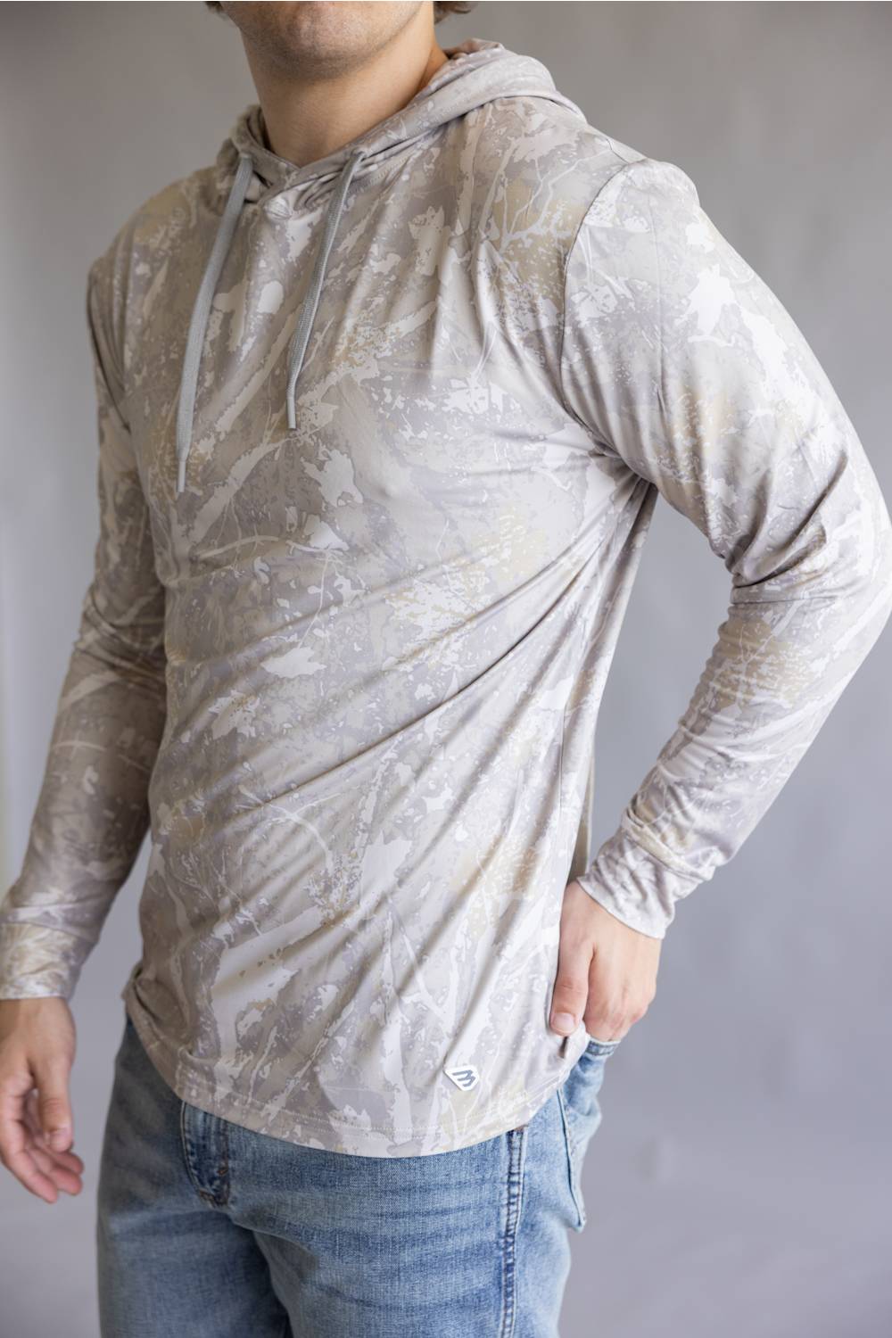 A man wearing a light beige and gray marbled long-sleeve hooded shirt with blue jeans, shown from the neck down against a neutral gray background.