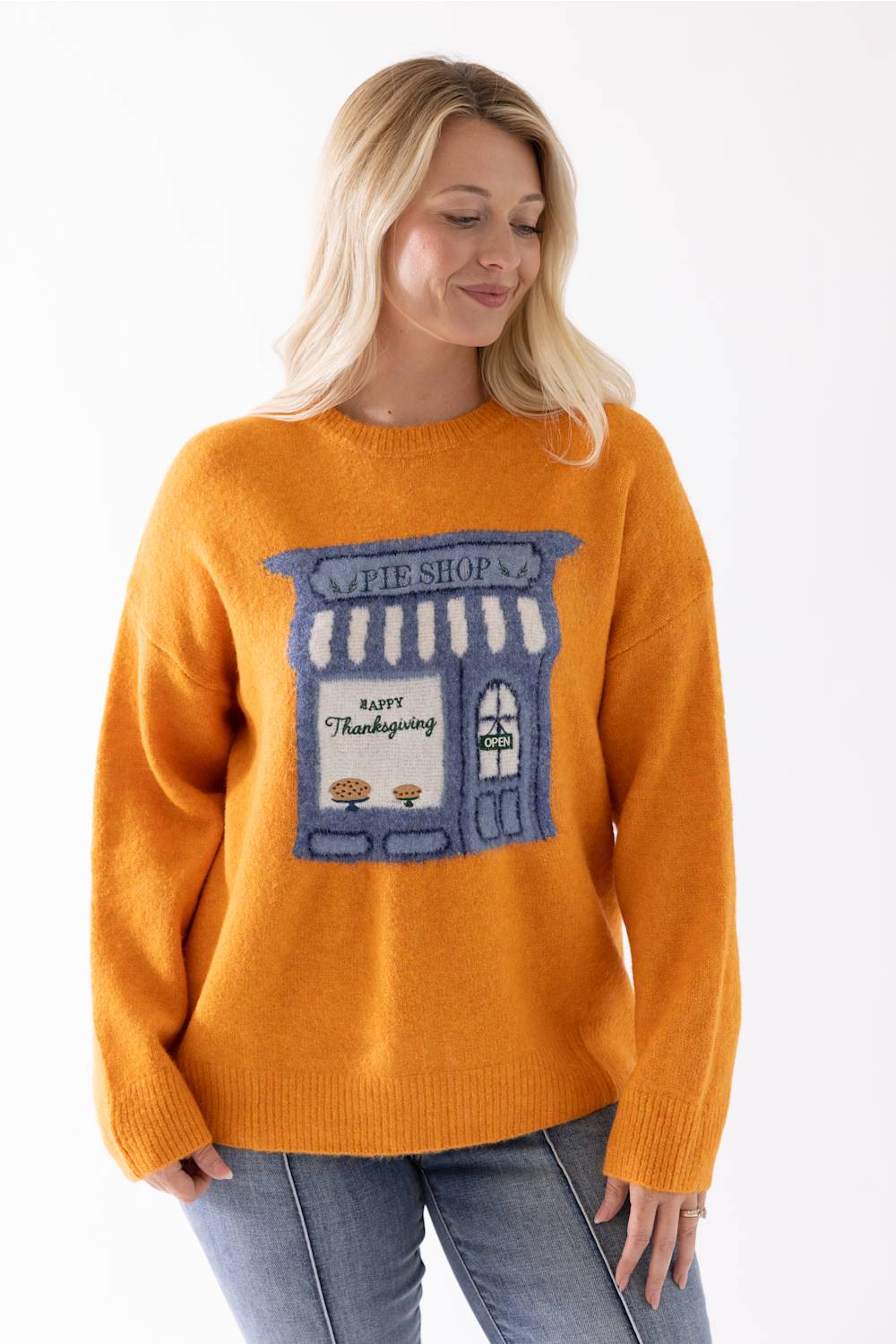 A woman wearing a bright orange crewneck sweater with a knitted graphic of a blue and white pie shop storefront that reads 'Happy Thanksgiving'.