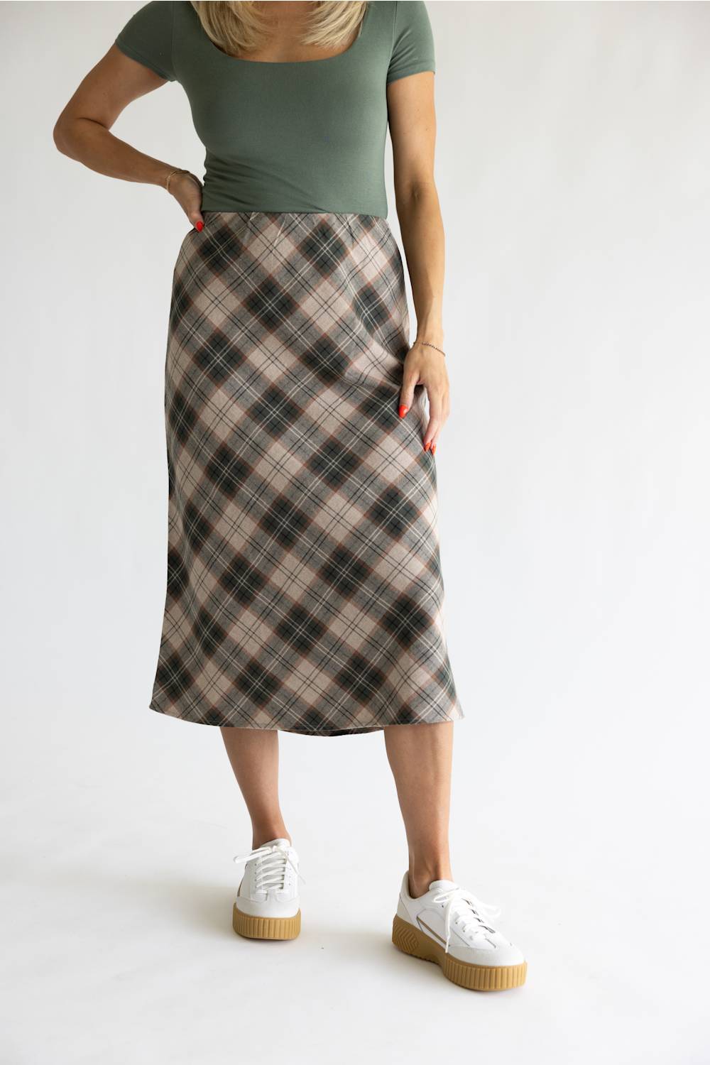 A woman modeling a knee-length plaid skirt in beige, brown, and black, paired with a muted green top and white platform sneakers, against a plain white studio background.