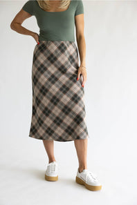 A woman modeling a knee-length plaid skirt in beige, brown, and black, paired with a muted green top and white platform sneakers, against a plain white studio background.