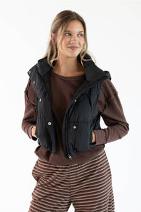 A woman modeling a black sleeveless puffer vest over a brown long-sleeve top and brown striped pants, photographed against a plain white studio background.
