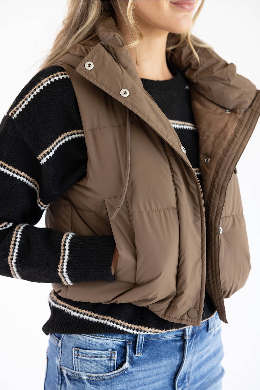 A woman wearing a brown puffer vest over a black and white striped knit sweater, paired with blue jeans, shown from the chest down against a plain white background.