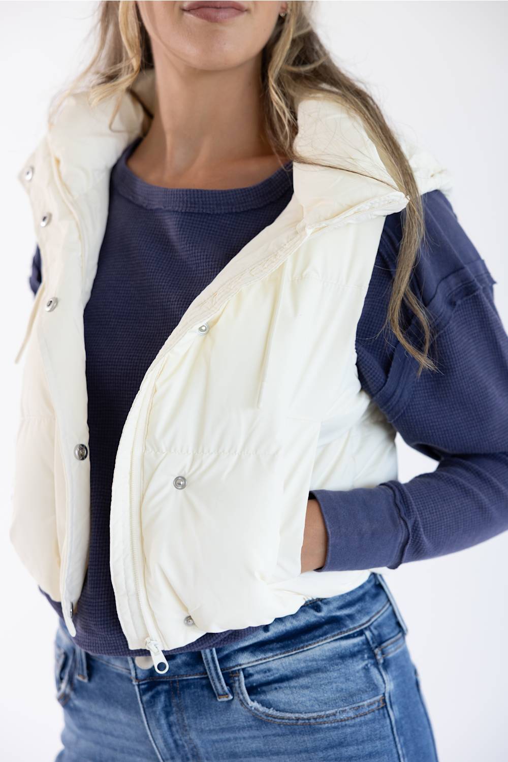 A woman wearing a cream-colored sleeveless puffer vest over a navy blue long-sleeve top and blue jeans, shown from the chin down against a plain white background.