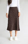 A woman wearing a dark brown midi skirt with a white polka dot pattern, styled with a white long-sleeve top and white sneakers against a light gray studio background.