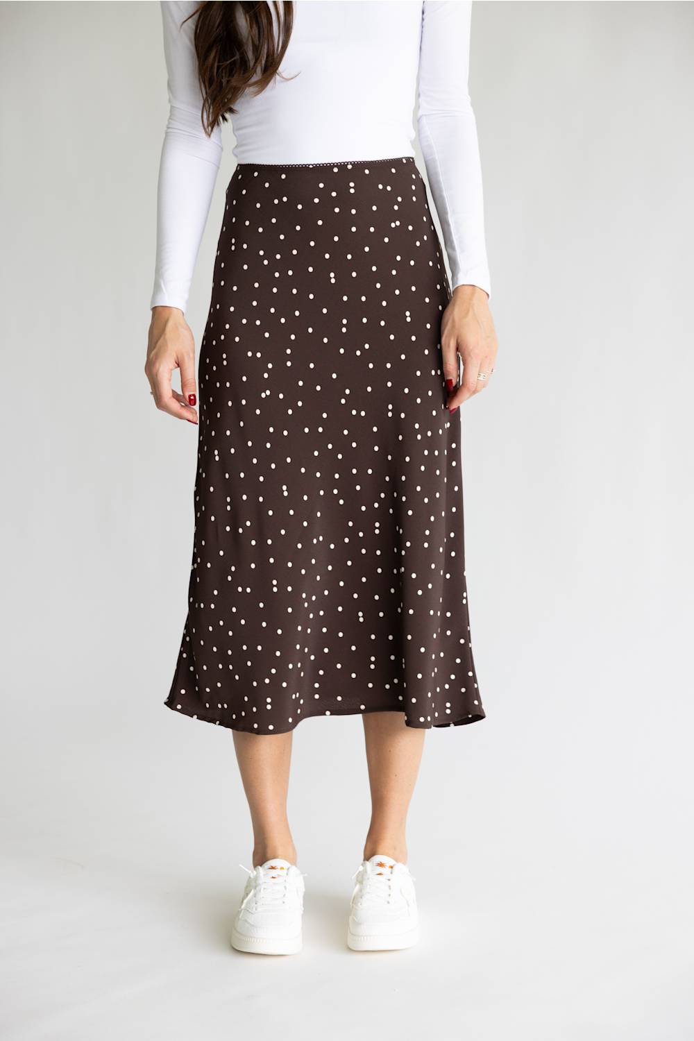A woman wearing a dark brown midi skirt with a white polka dot pattern, styled with a white long-sleeve top and white sneakers against a light gray studio background.