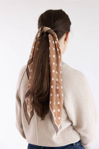 A woman viewed from behind wearing a beige scarf with white polka dots tied around her brown hair, paired with a cream knit sweater and blue jeans against a plain white background.