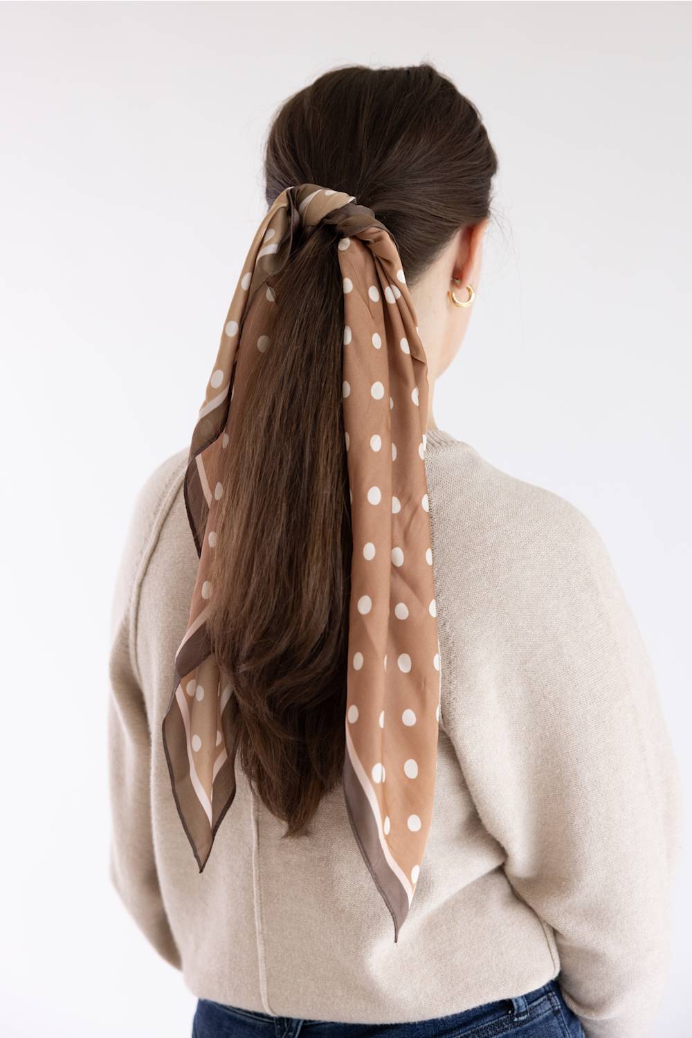 A woman viewed from behind wearing a beige scarf with white polka dots tied around her brown hair, paired with a cream knit sweater and blue jeans against a plain white background.