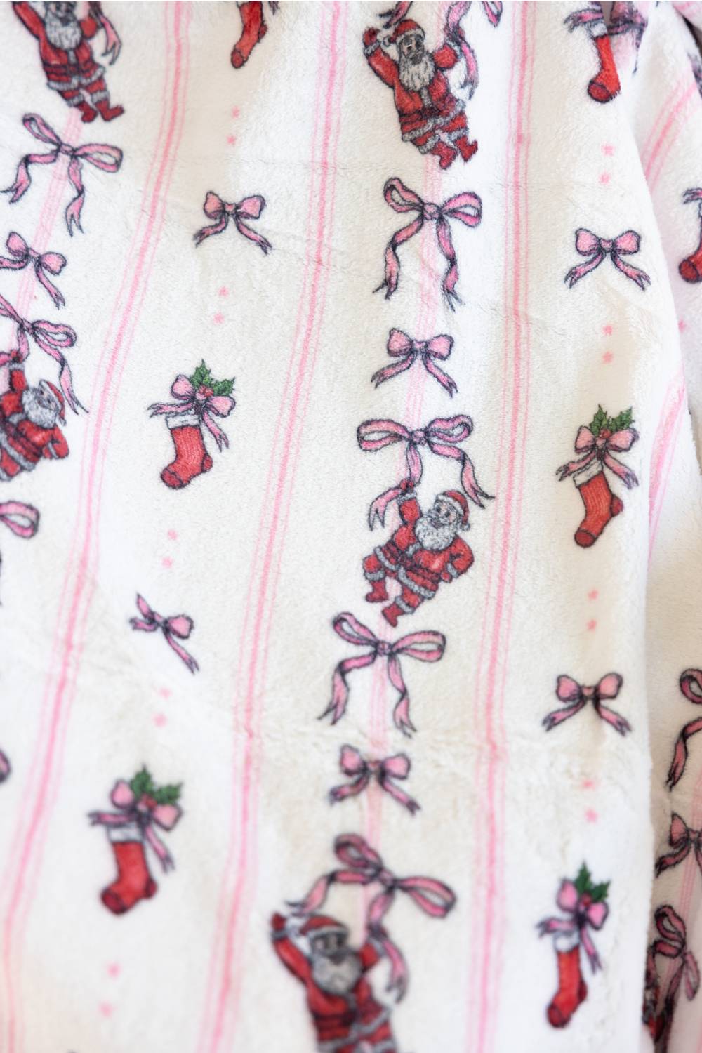 A close-up of a blanket with a pink striped pattern and a whimsical print of pink bows, red stocking and Santa Clause illustrations.