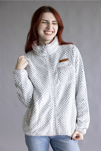 A woman with red hair wearing a white fleece jacket with a black micro-dot pattern and a brown chest patch, paired with blue jeans, posing against a solid gray studio background.