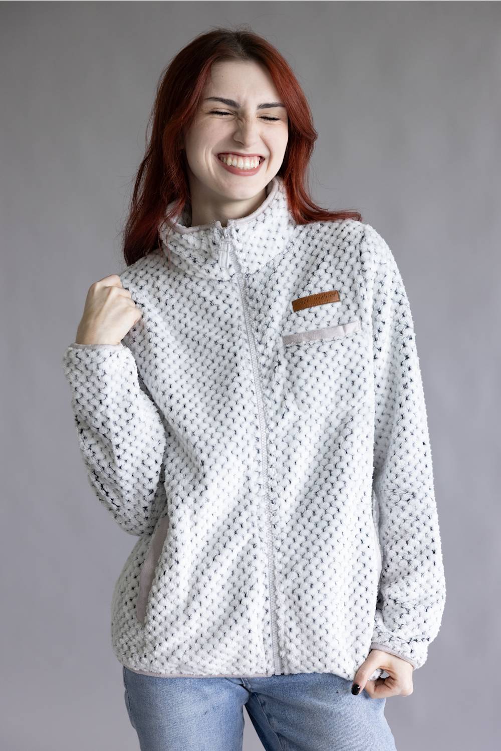 A woman with red hair wearing a white fleece jacket with a black micro-dot pattern and a brown chest patch, paired with blue jeans, posing against a solid gray studio background.