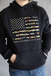 A person wearing a black pullover hoodie with a distressed American flag graphic on the chest. The flag stripes feature a camouflage pattern, and the text 'SIMPLY SOUTHERN OUTFITTERS' is printed below.