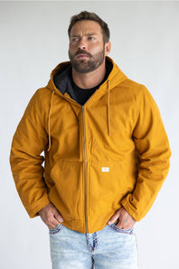 A man wearing a mustard yellow hooded jacket with a black interior lining and drawstrings, paired with light blue jeans, standing against a plain light gray background.