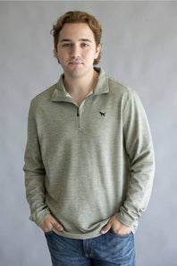 A young man with light brown hair wearing a light sage green heathered quarter-zip pullover sweater with a small black dog logo on the left chest, paired with blue jeans, standing against a neutral gray studio background.