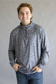 A smiling young man wearing a gray long-sleeve quarter-zip pullover with a subtle abstract pattern, paired with blue jeans, standing against a neutral gray studio background.