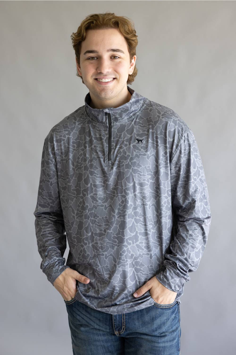 A smiling young man wearing a gray long-sleeve quarter-zip pullover with a subtle abstract pattern, paired with blue jeans, standing against a neutral gray studio background.