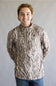 A smiling young man wearing a long-sleeve quarter-zip pullover jacket with a brown, beige, and gray bark-like camouflage pattern, paired with blue jeans, standing against a neutral gray studio background.